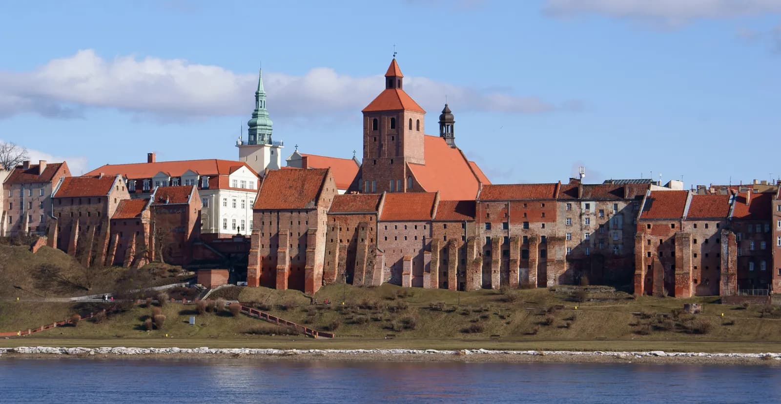 A scenic view of a historic European town situated on a hillside by a river, featuring a prominent church with a red roof and additional buildings with orange and brown roofs.